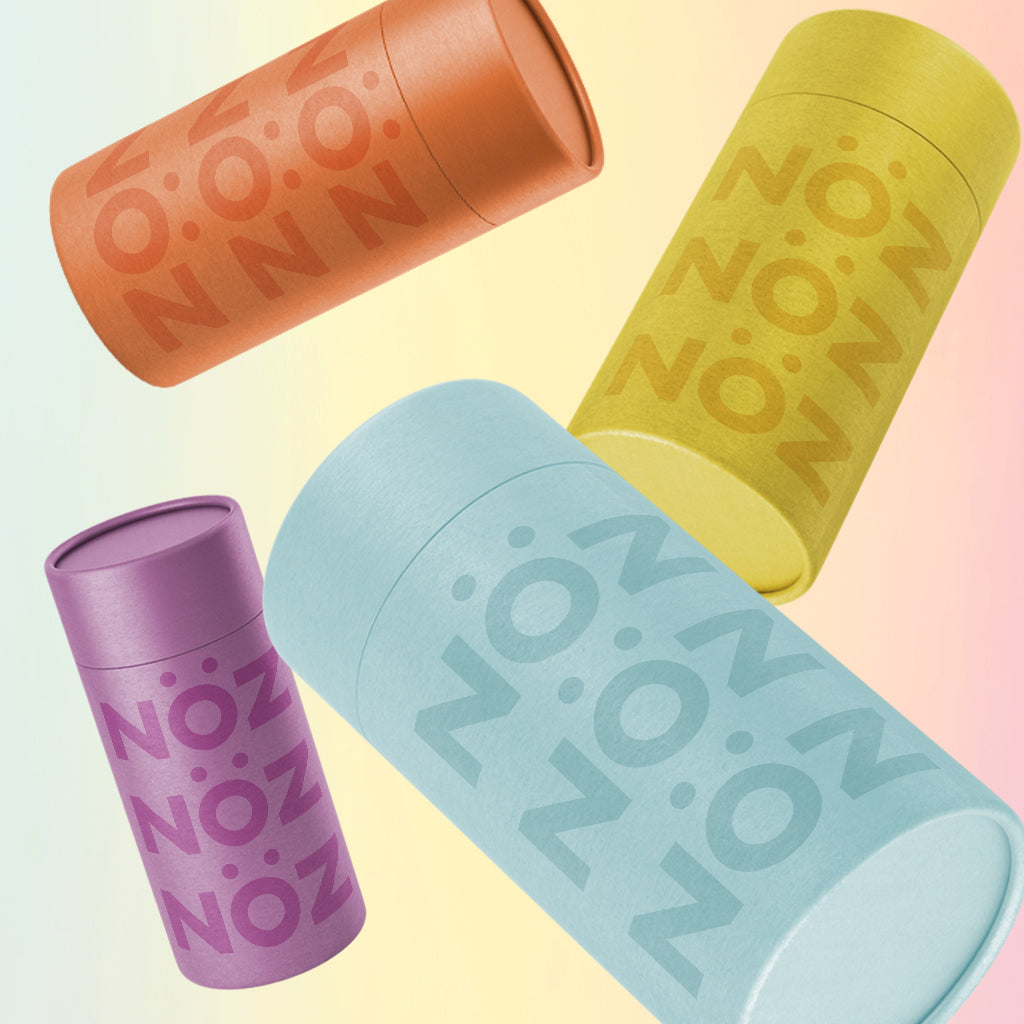 Four sticks of Nöz sunscreen in the shades Blue, Red, Yellow, and Purple against a pastel gradient background.